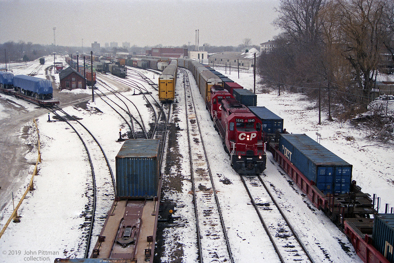 A pair of SD40-2's CP 5645 and CP5656 with a train of autoracks are facing east in this view of CP's Quebec Street Yard in London, taken from Quebec Street overpass.  The adjacent container train is single-stacked, otherwise it could not pass through the Detroit-Windsor tunnel. 
Over to the left under blue covers are two new Irish Rail (Iarnród Éireann) JT42HCW locomotives, units 232 and 230 on flatcars.  Ireland's wide rail gauge of 5' 3" prevents them travelling on their own wheels to their ship; their 3-axle trucks travel on another flatcar. 
Behind the pole to the right of tarped unit 232, the front of a locomotive coloured pale green can be seen. In this timeframe it is probably a new SD60i for Conrail, presumably in transit to them in primer.
Unfortunately the view west from this bridge is obstructed by cables that cross the tracks here.